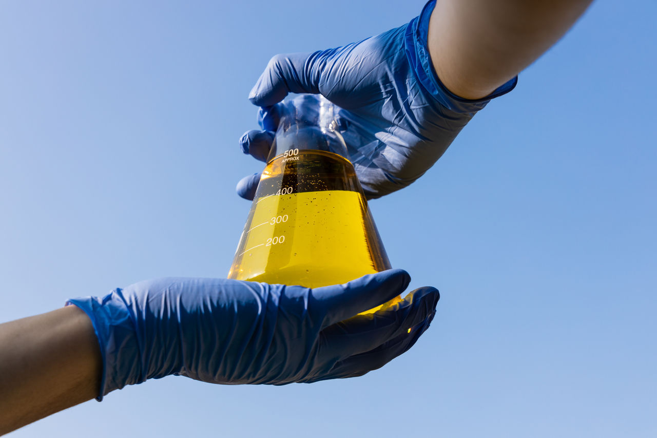 Hand with gloves holding beaker with ethanol biofuel against blue sky, observing its purity.